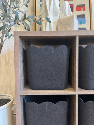 A set of three gray scalloped storage bins placed inside a wooden cube shelf, with a plant and various items in the background.