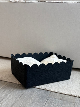 Black scalloped-edge basket with white towels on a wooden floor against a gray couch.